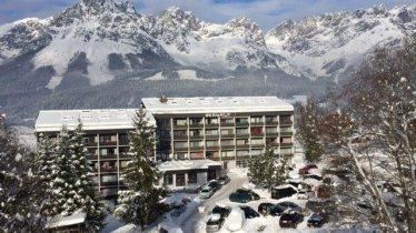 Ein modernes Hotel in einer schneebedeckten Landschaft. Im Hintergrund erheben sich majestätische Berge unter einem klaren Himmel.