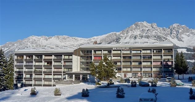 A modern hotel building, surrounded by snow-capped mountains and a clear blue sky. Winter is clearly visible with a white blanket of snow and green fir houses in the foreground.
