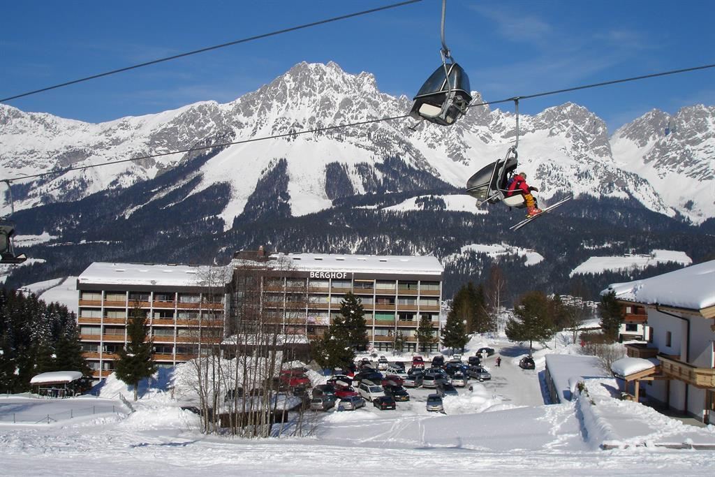 A large mountain landscape with snow-covered peaks and a ski lift in the foreground. In the background, a hotel and a parking facility are visible.
