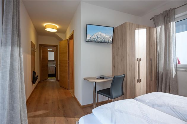 A bright room with two single beds, a desk, and a TV on the wall. The wood paneling and curtains create a cozy atmosphere.