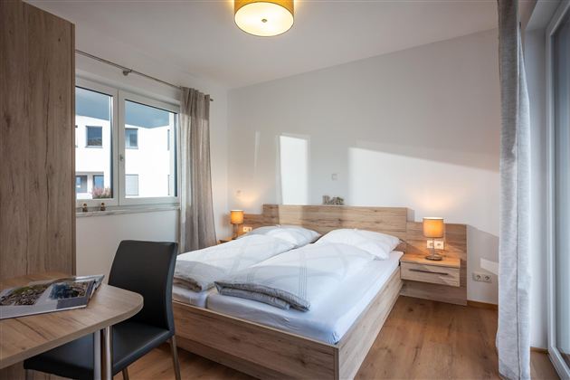 A modern bedroom with a double bed and wooden furniture. The room is bright and has large windows for natural light.