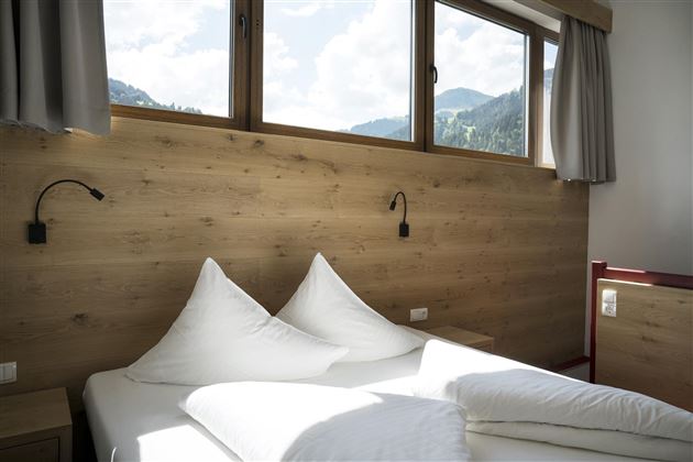 Ein helles Schlafzimmer mit Holzpaneelen und großen Fenstern. Zwei große, weiße Kissen liegen auf dem Bett, während die schönen Berglandschaften sichtbar sind.