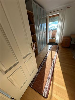 Ein heller Raum mit einem Balkon und viel Tageslicht. Zu sehen sind ein Teppich, ein Schrank und eine helle Holzverarbeitung.