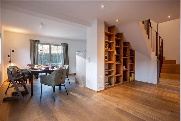 Ein modernes Wohnzimmer mit einem Essbereich und einer Treppe im Hintergrund. Holzboden und große Fenster sorgen für eine helle und einladende Atmosphäre.