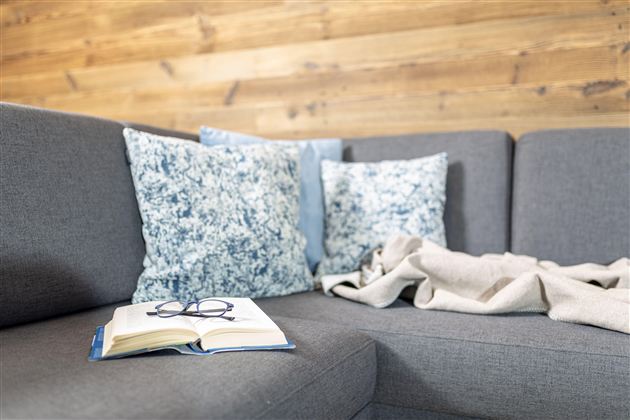 A cozy living room with a gray sofa, colorful cushions, and a blanket. On the table lies an open book and a pair of glasses.