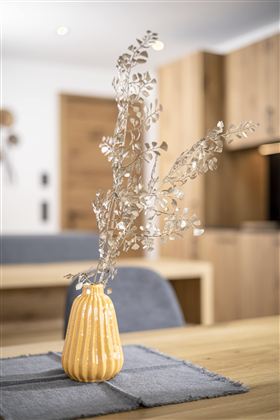 A yellow vase with silver branches stands on a wooden table. In the background, modern furniture and a neutral color palette are visible.