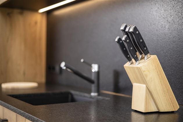 A modern kitchen environment with a wooden cutting block holding five sharp knives. In the background, a sink and a faucet are visible.