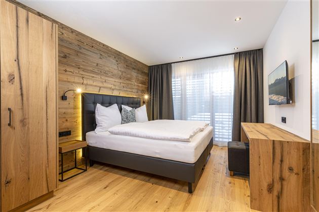 A modern bedroom with wooden paneling and a large window. It is stylishly furnished with a cozy bed and a wooden table.