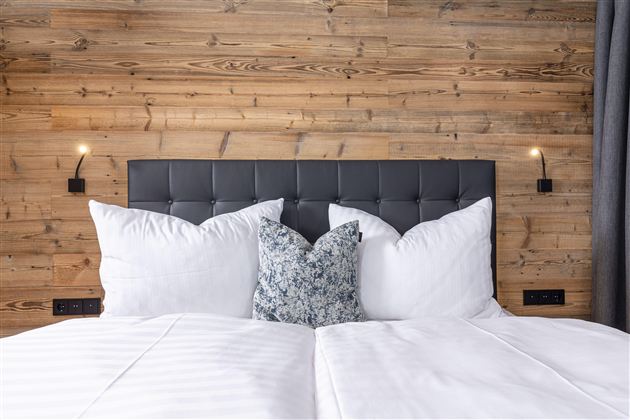 A cozy bedroom with a modern headboard and soft pillows. The wooden wall gives the room a warm atmosphere.