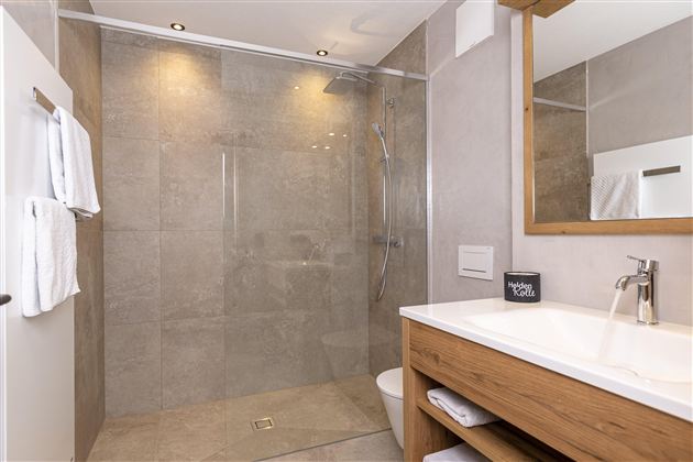A modern bathroom with a large shower and gray tiles. The wash area has a wooden cabinet and a mirror.