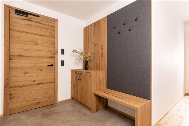 A modern entrance area with a wooden door and a stylish wooden bench. On the wall, there is a gray area with hooks and a vase with dried flowers.