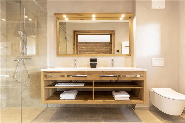 A modern bathroom with a large mirror wall and an elegant wooden washstand. The shower is made of glass and there are fresh towels on the open shelf.