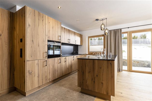 Eine moderne Küche mit Holzschränken und eleganten Beleuchtungselementen. Große Fenster sorgen für viel Tageslicht und einen Blick nach draußen.