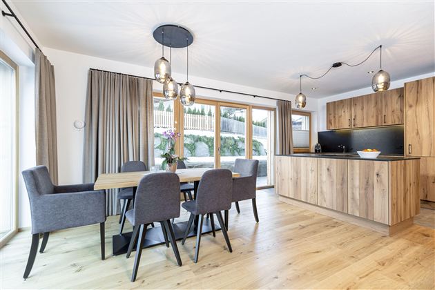 Eine moderne Küche mit einem Essbereich aus Holz und stilvollen Stühlen. Große Fenster lassen viel Tageslicht herein und bieten einen Blick auf die Außenterrasse.