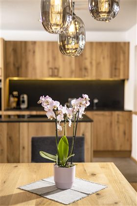 An elegant table with a blooming orchid in a pot. In the background, there is a modern kitchen with wooden cabinets and bright lighting.