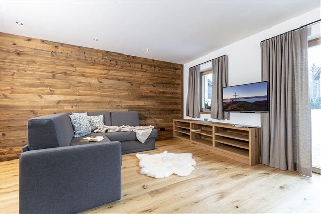 A modern living room with a gray sofa and wooden walls. A television is placed on a wooden cabinet and large windows let in plenty of light.