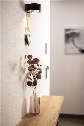 A stylish interior with a wooden console and a pink vase filled with dried branches. A modern lamp hangs on the wall along with a heart-shaped decorative piece.