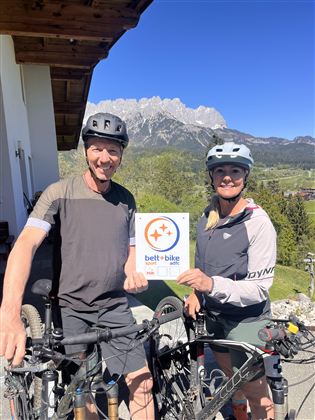 A pair of cyclists stands next to their bicycles holding a sign with the logo "bett+bike". In the background, mountains and a clear blue sky can be seen.