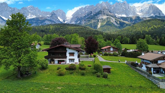 A picturesque landscape with a traditional house and majestic mountains in the background.  
Surrounded by green meadows and trees, the scene radiates tranquility and natural beauty.