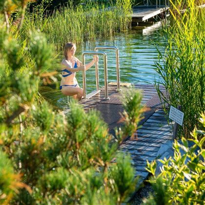 A woman in a swimsuit stands on a steps leading to a body of water. In the background, tall plants and a pier can be seen.