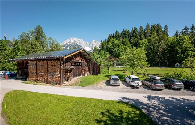 Een rustiek gebouw omringd door groene weiden en bomen. Op de achtergrond zijn sneeuwbedekte bergen te zien.
