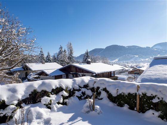 A winter landscape with snow-covered roofs and trees. In the background, gentle mountains and a clear blue sky can be seen.
