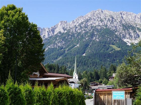 Een schilderachtig Berglandschap met hoge toppen en weelderig groene bossen. Op de voorgrond zijn houten huizen en een kerk met een spitse toren te zien.