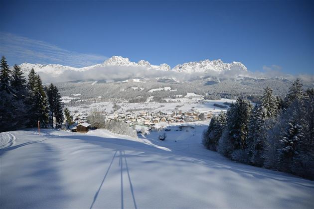 Eine schneebedeckte Landschaft mit Bergen im Hintergrund und einem kleinen Dorf im Tal. Der Himmel ist klar und blau, was eine friedliche Winteratmosphäre schafft.