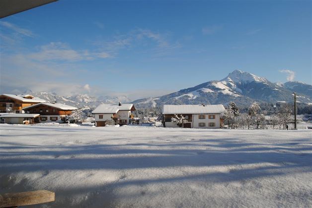 A winter landscape with snow-covered huts and mountains in the background. The sky is clear and blue.