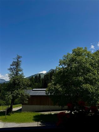 A clear blue sky over a picturesque landscape with green trees. In the foreground, there is a modern wooden building.