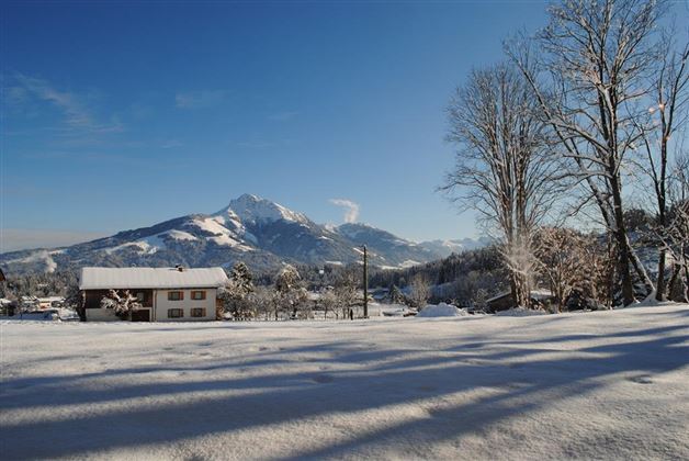 A winter landscape with snow-covered fields and a view of snow-covered mountains. In the foreground, there is a cozy house under a clear blue sky.