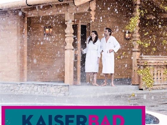 A couple in bathrobes stands in front of a wooden cabin and enjoys the snow. The atmosphere is relaxed and inviting.