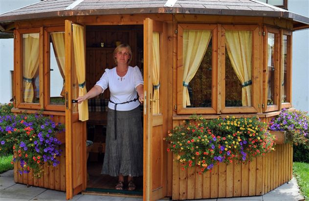 A woman stands in the open door of a cozy wooden house. Flowers adorn the windows, creating an inviting atmosphere.