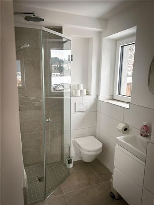 Een moderne badkamer met een glazen douche en een wit toilet. Er is een raam dat licht binnenlaat, en een kleine wastafelonderkast.
