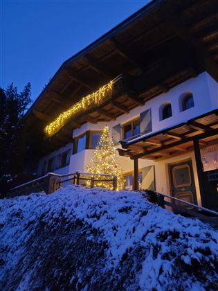 Een gezellig huis in de winter met sneeuw en feestelijke verlichting. Een kerstboom staat voor de voordeur en zorgt voor een uitnodigende sfeer.