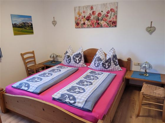 Een uitnodigende slaapkamer met een groot tweepersoonsbed en kleurrijke kussens. Aan de muur hangt een kunstwerk met bloemenmotieven.
