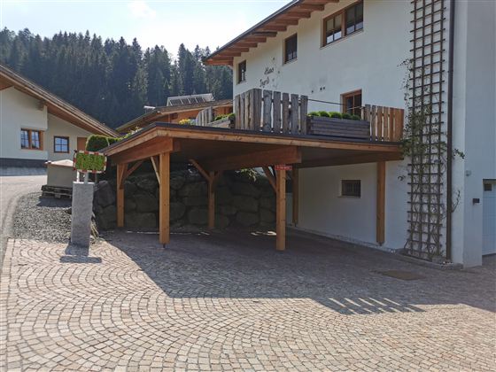 Ein modernes Haus mit einer überdachten Terrasse und einem gepflasterten Parkplatz. Im Hintergrund sind grüne Bäume und eine ruhige Landschaft sichtbar.