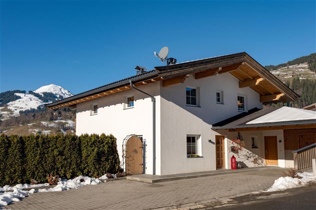 Ein modernes Haus mit einem Holzdach und einer hellen Fassade. Im Hintergrund sind verschneite Berge zu sehen.
