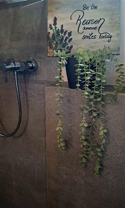 Een moderne douche met een muur van beton en een kraan. Boven hangt een bord met de tekst "Be the reason someone smiles today" en een plant in een pot.