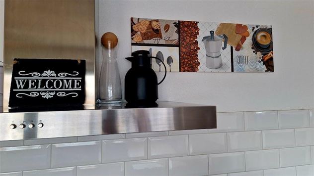 Een moderne keuken met een elegante metalen afdekking. Op het aanrecht staan een glazen karaf, een zwarte koffiecontainer en een decoratieve wandversiering met koffie- en koekjesafbeeldingen.