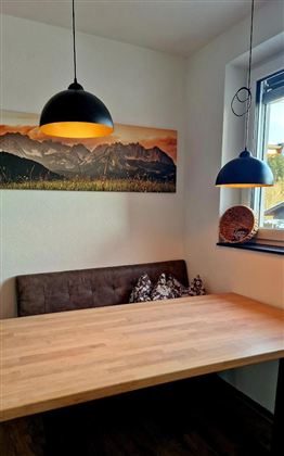 Een gezellige eetruimte met een houten tafel en een bank. Boven de tafel hangen twee stijlvolle lampen en aan de muur hangt een schilderij van bergen.