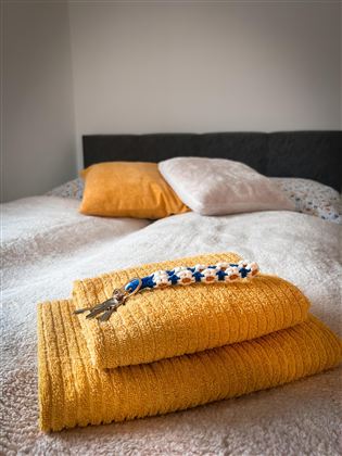 Een gezellig bed met oranje kussens en verse, gele handdoeken. Aan de handdoek hangt een gekleurde sleutelhanger.