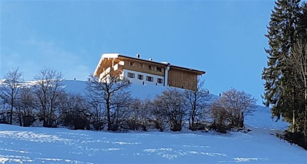 A beautiful house that stands on a snow-covered hill. The sky is clear and blue, with scattered trees in the foreground.