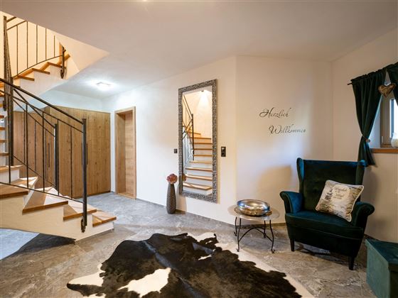 Ein einladender Eingangsbereich mit einer schicken Treppe und einem grünen Sessel. Auf dem Boden liegt ein Kuhfell, und eine Wand mit der Aufschrift „Herzlich Willkommen“ sorgt für eine freundliche Atmosphäre.