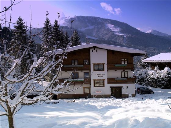 Ein gemütliches Chalet im Schnee, umgeben von schneebedeckten Bäumen. Im Hintergrund sind majestätische Berge und ein klarer Himmel zu sehen.