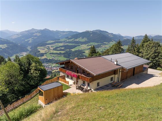 Ein modernes Holzhaus auf einer Anhöhe mit Blick auf die umliegenden Berge. Die Landschaft ist grün und hügelig, unter einem klaren blauen Himmel.