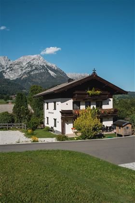 Ein traditionelles chalet mit braunem Dach und bunten Blumen. Im Hintergrund sind majestätische Berge und ein klarer blauer Himmel zu sehen.