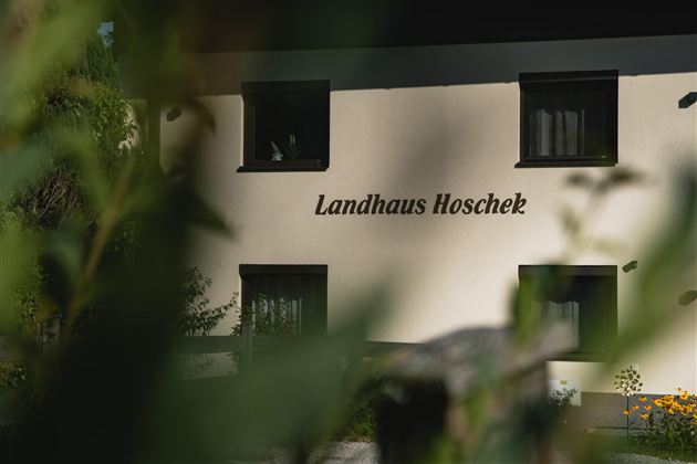 Ein charmantes Landhaus mit der Aufschrift "Landhaus Hoschek". Es ist von Pflanzen umgeben und strahlt eine einladende Atmosphäre aus.