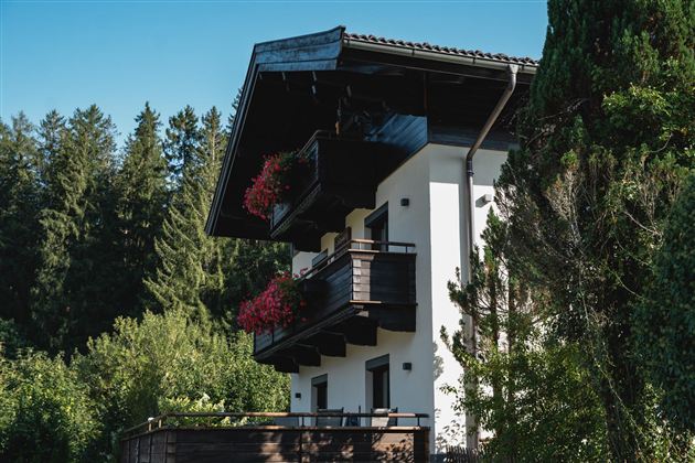 Ein modernes Haus umgeben von Bäumen und Grün. Der Balkon ist mit Blumen geschmückt und die Fassade zeigt einen stilvollen Kontrast zwischen Holz und Putz.