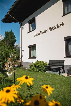 Ein charmantes Landhaus mit dem Namen "Landhaus Hoschek" und einem schönen Garten. Sonnenblumen blühen im Vordergrund und schaffen eine einladende Atmosphäre.